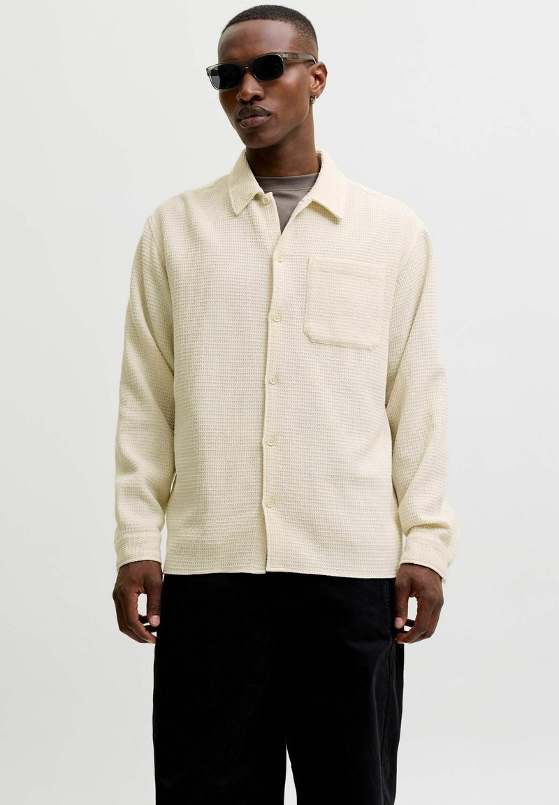 Homme portant des lunettes de soleil noires, une chemise crème texturée boutonnée avec une poche poitrine, et un pantalon noir sur un fond clair uni.