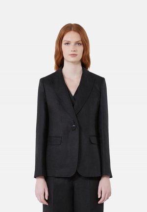 Giovane donna con capelli rossi che indossa una giacca nera aderente e pantaloni abbinati, in piedi davanti a uno sfondo grigio chiaro uniforme.
