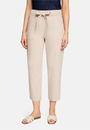 Femme portant un pantalon beige court avec poches avant et taille nouée, associée à des sandales plates crème et un haut bleu foncé.