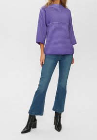 Frau in einem lila gestreiften Pullover, blauen Schlagjeans und schwarzen High Heels, die vor einem schlichten weißen Hintergrund steht.
