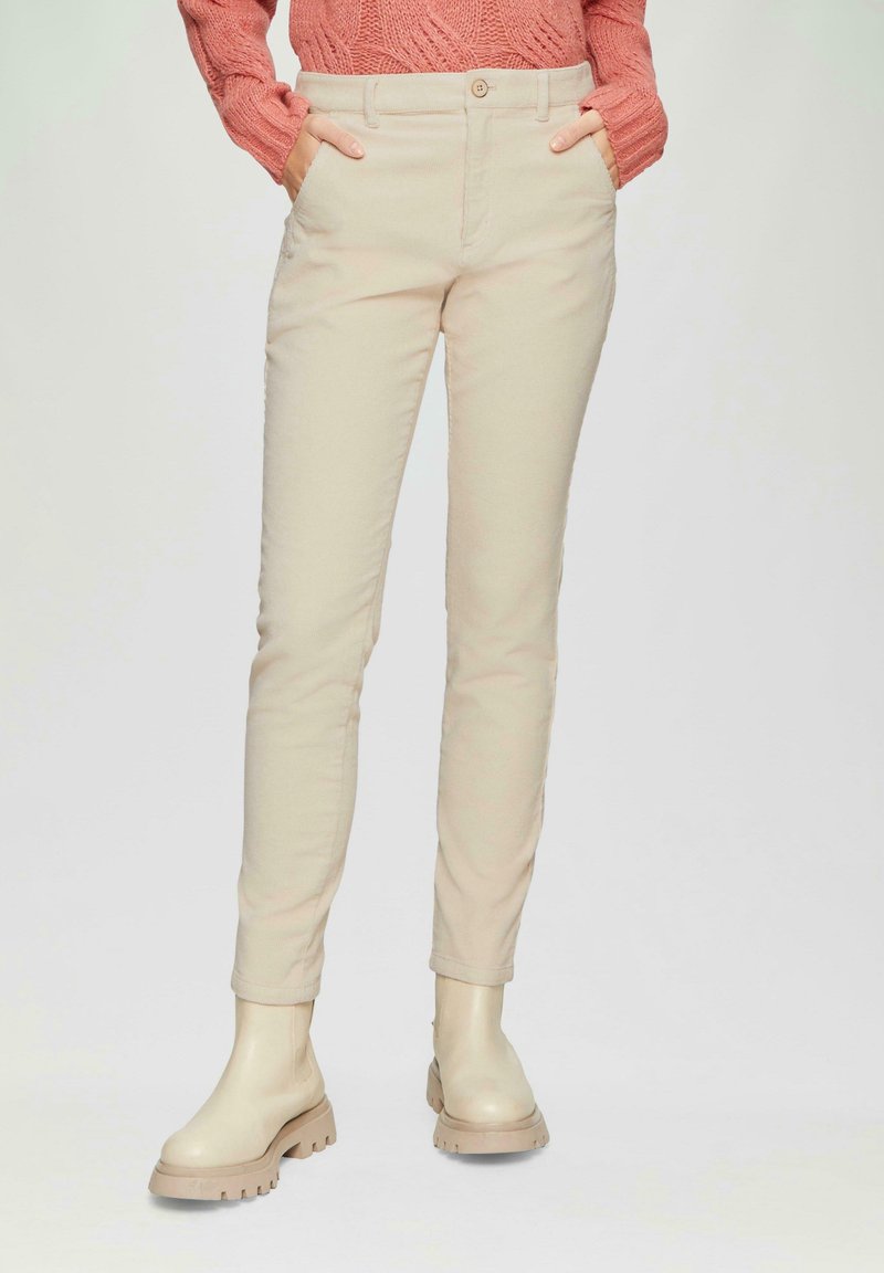 Personne portant un pantalon beige slim, des bottines beiges épaisses et un pull corail texturé, les mains dans les poches.