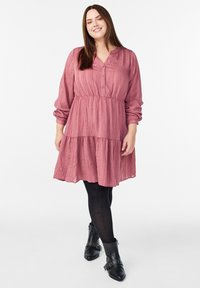 Vestido texturizado color rosa con tapeta de botones, cintura fruncida y falda en capas. Llevado con leggings negros y botas de tobillo.