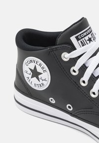 Adidași negri tip gheată cu șireturi albe, având un patch rotund cu logo, ochiuri metalice argintii și o talpă cu dungi albe. Material sintetic neted.