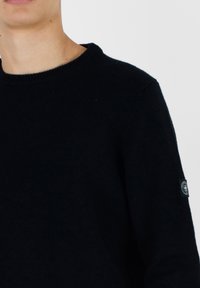 Maglione blu scuro con scollatura tonda e polsini a costine. Presenta una piccola etichetta in tessuto sul braccio sinistro. Texture morbida, design lavorato a maglia.