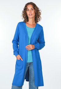 Blauer strukturierten Cardigan mit langem, offenem Design, ausgestattet mit zwei vorderen Taschen. Getragen über einem gestreiften, kurzärmeligen Oberteil und einer Jeans.