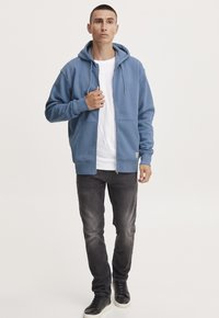 Sudadera azul con cremallera y bolsillos frontales, con cordón ajustable, encima de una camiseta blanca, combinada con vaqueros oscuros y zapatillas negras.