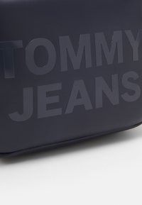 Borsa rettangolare blu navy con lettering in rilievo "TOMMY JEANS". Texture liscia e opaca, realizzata in materiale sintetico. Design compatto.
