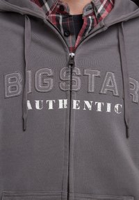 Grå zip-up hættetrøje lavet af en bomuldsblanding, med et frontprint "BIG STAR AUTHENTIC" i hvidt og omkranset af mørkegrå. Tekstureret stof, snore.