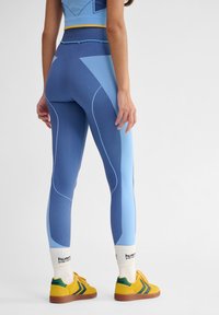 Höga leggings i blå nyanser med kontrasterande ljusblå paneler och räfflad textur, kombinerade med gula sneakers och vita strumpor.