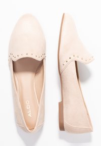 Chaussures slip-on beiges avec une finition en suède texturé, bout arrondi et bord festonné orné de clous en or décoratifs. Talon plat.