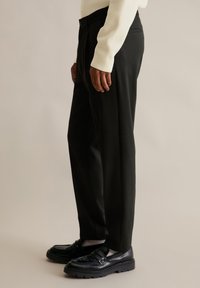 Person wearing black tailored trousers, white socks, black leather loafers, and a cream-colored long-sleeve top, standing sideways on beige background.