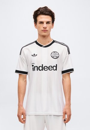 Giovane uomo che indossa la maglia da calcio bianca dell'Eintracht Francoforte con rifiniture nere, logo Adidas e sponsor Indeed, in piedi su sfondo semplice.