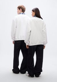 Λευκά φαρδιά φούτερ με χαμηλωμένους ώμους, σε συνδυασμό με άνετα σκούρα τζιν. Εντυπωσιακές πολύχρωμες λεπτομέρειες κοντά στο τελείωμα των φούτερ.