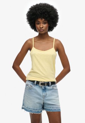 Débardeur jaune à fines bretelles, associé à un short en denim bleu clair avec un ourlet effiloché et une ceinture noire avec une boucle en argent.