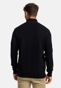 Marinblå stickad tröja med en fiskbensmönster, hög krage och ribbade mudd och nederkant. Tyget verkar strukturerat och varmt.