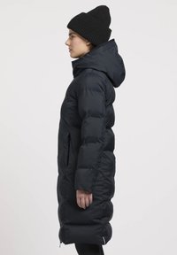 Čierna nafúknutá bunda s kapucňou, s elegantným dizajnom, prešívanou textúrou a bočnými zipsovými vreckami. Nosená s čiernym pleteným čiapkou.