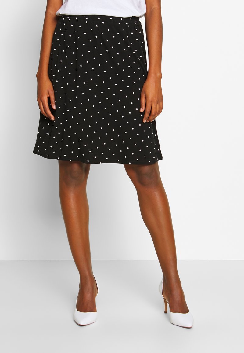 Femme portant une jupe noire à pois blancs longueur genoux et des escarpins blancs, debout devant un fond uni.