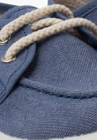 Baskets en toile bleue avec des lacets tressés beiges, œillets en acier inoxydable et semelle en caoutchouc blanche. Présente des détails cousus et un tissu texturé.