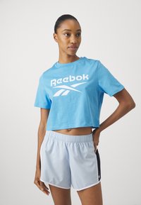 Lyseblå cropped t-skjorte med hvit "Reebok" logo, kombinert med lysegrå treningsshorts med elastisk liv og sidelommer.