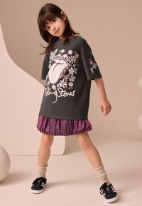 Grå oversize T-shirt med Rolling Stones-logotyp och blommiga detaljer, kombinerad med en lila kjol med veck. Svarta sneakers och krämfärgade strumpor fullbordar looken.