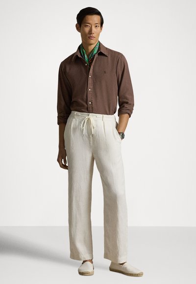 Καφέ πουκάμισο με κουμπιά, λευκό παντελόνι από λινό με κορδόνι και μπεζ παπούτσια τύπου slip-on. Πράσινο φουλάρι με σχέδια προσθέτει χρώμα στην εμφάνιση.
