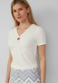 Witte korte mouwen top met een V-hals en een centrale sleutelgatdetail, gemaakt van zachte stof met een gladde textuur, gecombineerd met een patroon rok.