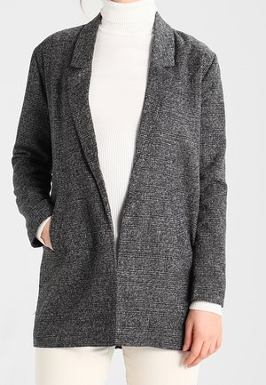 Kvinde iført en grå tekstureret blazer over en hvid rullekravesweater og lyse bukser, med den ene hånd i blazerens lomme.