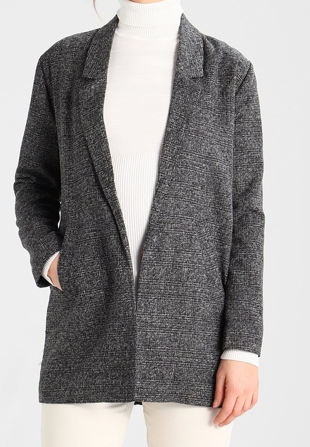 Femme portant un blazer gris texturé par-dessus un pull à col roulé blanc et un pantalon clair, avec une main dans la poche du blazer.