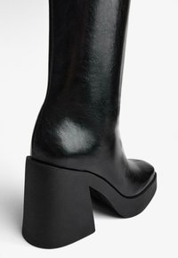 Botă de genunchi neagră, confecționată din piele lucioasă, cu un toc gros de culoare neagră. Prezintă o textură netedă și o siluetă eleganță.