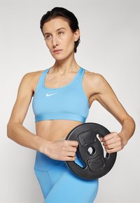 Lyseblå sports-bh med Nike-logo, parret med matchende tights. Holder en sort vægtskive, der viser et glat, moderne design.