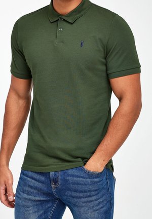 Homme portant un polo vert foncé avec un logo brodé bleu marine sur la poitrine gauche et un jean bleu, la main droite dans la poche.