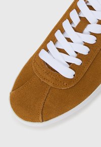 Close-up de uma bota desportiva em camurça bege com atacadores brancos e detalhes de costura sobre um fundo branco.