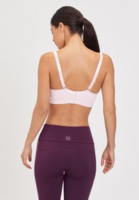 Femme aux cheveux foncés attachés en queue de cheval, portant un soutien-gorge rose clair et un legging taille haute violet, vue de dos sur un fond uni.