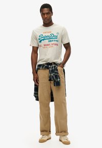 Lichtgrijze t-shirt met vintage graphics, gecombineerd met lichtbruine ribfluwelen broek en een zwart/groen geruite blouse die om de taille is geknoopt. Lichte sneakers.