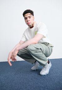 Ανοιχτόχρωμο μπλουζάκι με πράσινη γραφική παράσταση, ελαιοπράσινα cargo παντελόνια και γκρι αθλητικά παπούτσια με κόκκινες λεπτομέρειες, τοποθετημένα σε ανοιχτό φόντο.