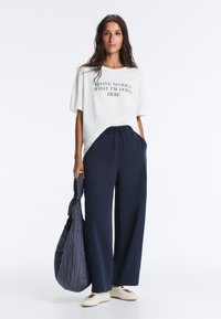 Femme aux longs cheveux foncés portant un t-shirt blanc avec l'inscription « Je n'ai aucune idée de ce que je fais ici », pantalon large bleu marine, chaussures blanches, tenant un sac rayé.