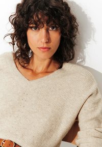 Pull en maille beige avec un col en V, tissu texturé et détails subtils. Modèle portant une ceinture en cuir marron, arrière-plan blanc uni.