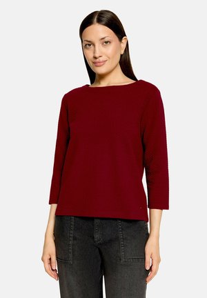 Femme aux cheveux raides et foncés portant un haut à manches longues rouge foncé et un jean noir, debout devant un fond clair uni.