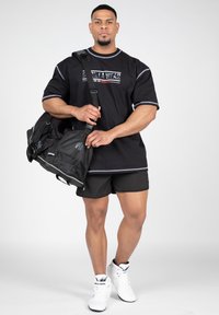 Fekete sportpóló szürke részletekkel, fekete rövidnadrág és fehér magasszárú sportcipő. Kézben tart egy fekete sporttáskát, hálós díszítéssel.