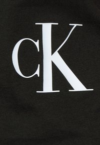 Fabric black con una textura suave que presenta un gran logo blanco "CK" centrado en la prenda. Diseño simple y audaz con alto contraste.