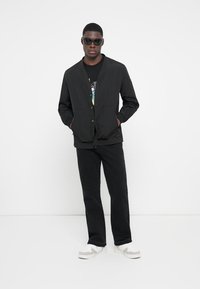 Sort let bomberjakke med trykknapper, parret med sorte jeans og hvide sneakers. Modellen har solbriller på og en t-shirt med grafik.