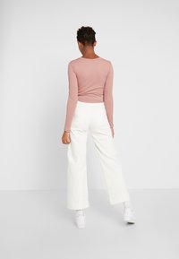 Růžový žebrovaný top s dlouhým rukávem a přiléhavým střihem, doplněný vysokými širokými bílými kalhotami. Modelka má na nohou bílé tenisky. Jednoduchý a čistý design.