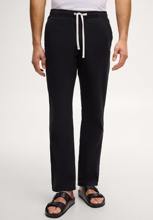 Homme portant un pantalon noir à cordon et des sandales noires à boucle, debout devant un fond clair uni.