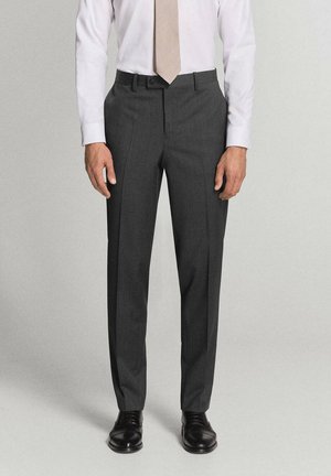 Hombre con pantalones de vestir gris oscuro, camisa blanca, corbata beige y zapatos de vestir negros, de pie frente a un fondo liso.