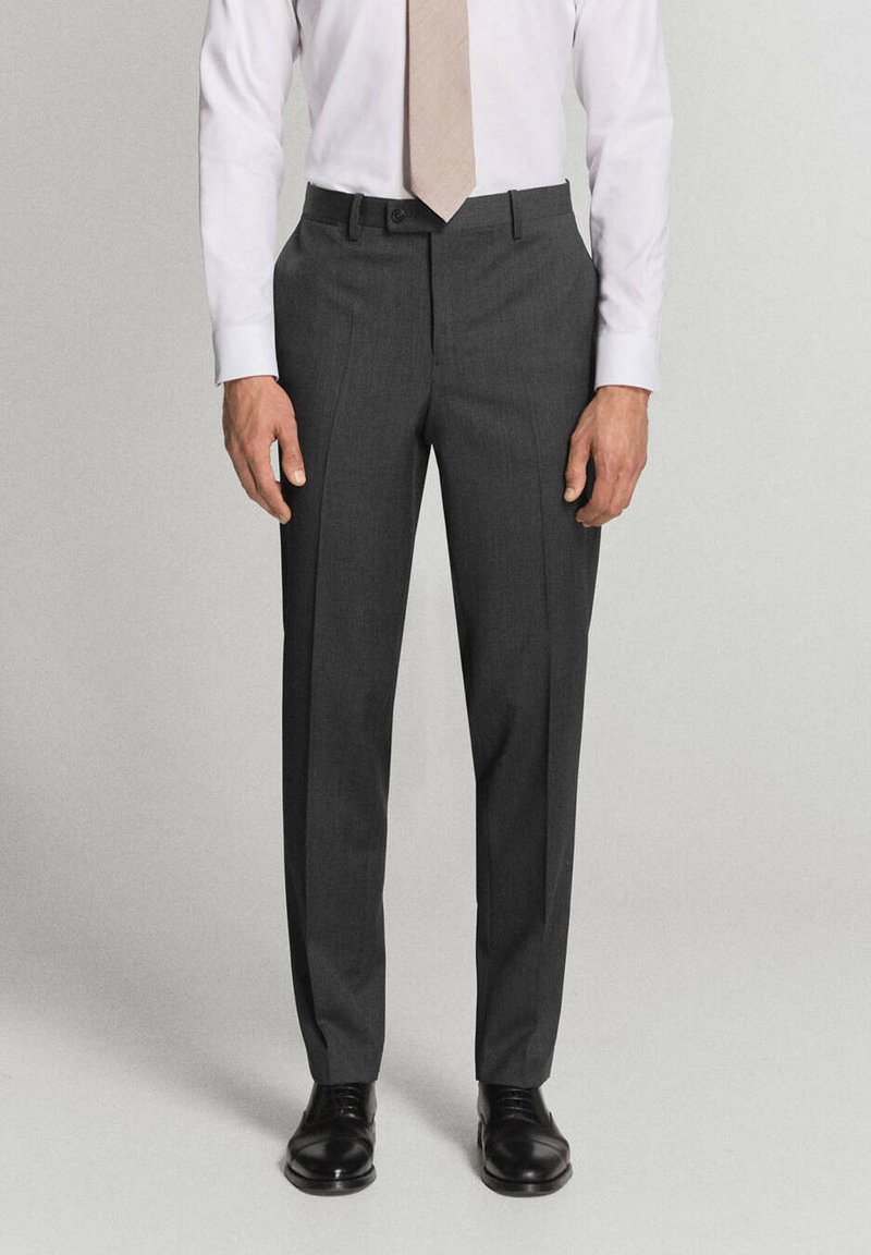 Homme portant un pantalon de costume gris foncé, une chemise blanche, une cravate beige et des chaussures noires, debout devant un fond uni.
