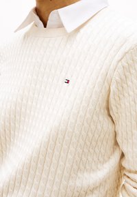 Homme portant un pull en maille torsadée blanc par-dessus une chemise blanche à col avec un petit logo Tommy Hilfiger rouge, blanc et bleu sur la poitrine.