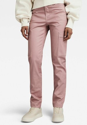 Osoba ubrana w jasnoróżowe, proste spodnie cargo z kieszeniami bocznymi, kremową bluzę i beżowe tenisówki, stojąca na białym tle.