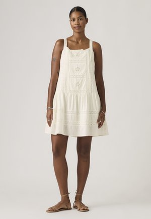 Femme portant une robe sans manches, de couleur crème, longue jusqu’au genou avec des détails en dentelle, debout pieds nus devant un fond clair uni.