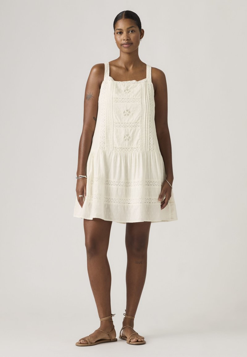 Γυναίκα που φοράει μαν sleeveless, κρεμ φόρεμα μήκους έως το γόνατο με δαντελένιες λεπτομέρειες, όρθια ξυπόλυτη σε ένα απλό φωτεινό φόντο.