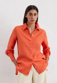 Camicia button-up in lino arancione con colletto classico, maniche lunghe e taschino sul petto. Il tessuto presenta una leggera texture.